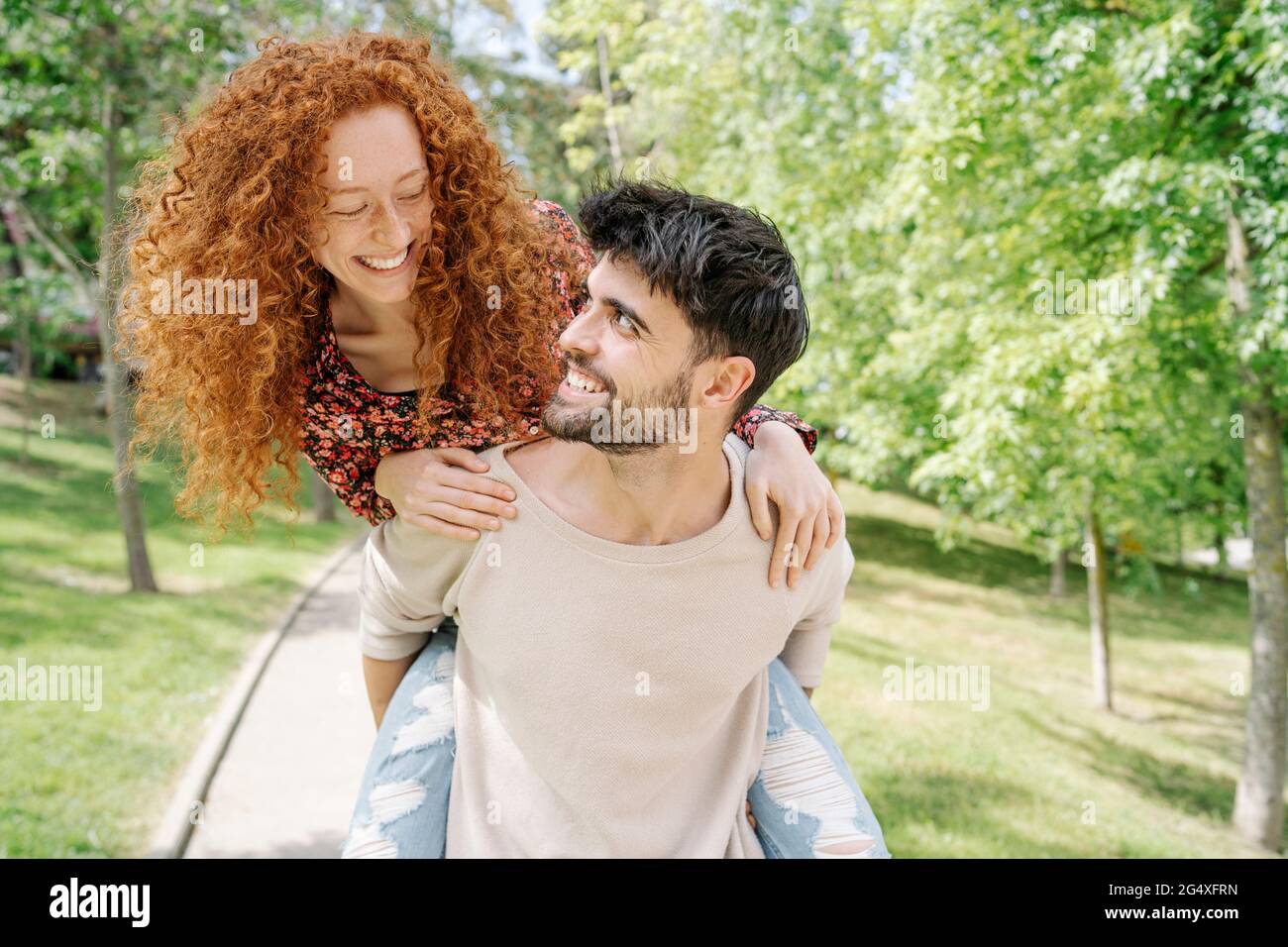 Novio sonriente llevando novia piggy de vuelta en el parque público Foto de stock