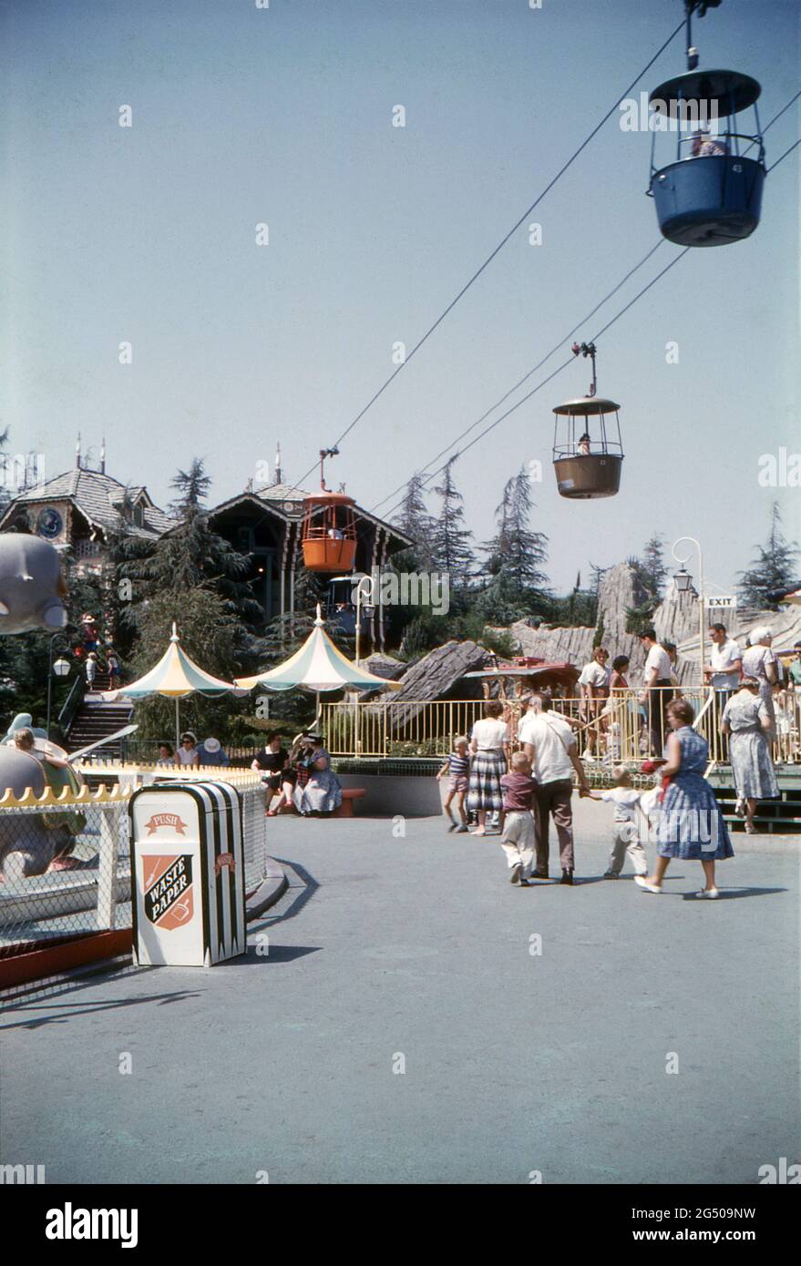 Disneyland, California, 1959. Vistas a la terminal del teleférico Swiss Chalet Skyway en Fantasyland. A la izquierda está el paseo Dumbo Flying Elephants y a la derecha está la salida para el Casey Jr. Circus Train. Los visitantes pasan y se refugian bajo las sombrillas. Foto de stock