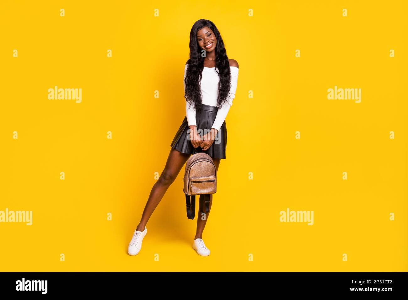 Cuerpo largo tamaño foto morena pelado negro sonriente sosteniendo una mochila con falda corta aislada sobre fondo de color amarillo vibrante Foto de stock