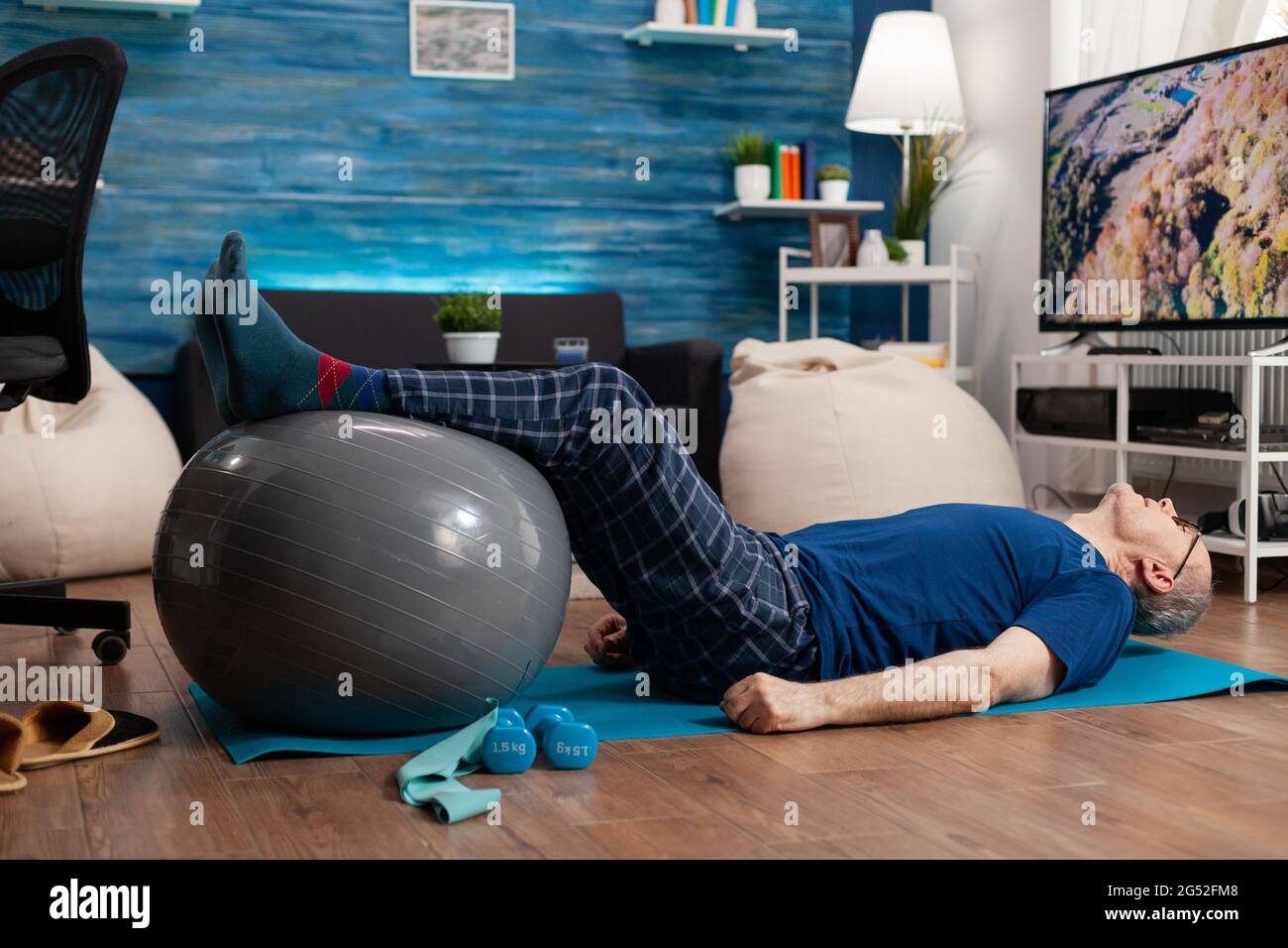 Jubilado hombre mayor haciendo bienestar calentar piernas con pelota suiza sentado en colchoneta de yoga en la sala de estar. Jubilado pensionista que practica ejercicios abdominales trabajando en la salud muscular del cuerpo Foto de stock