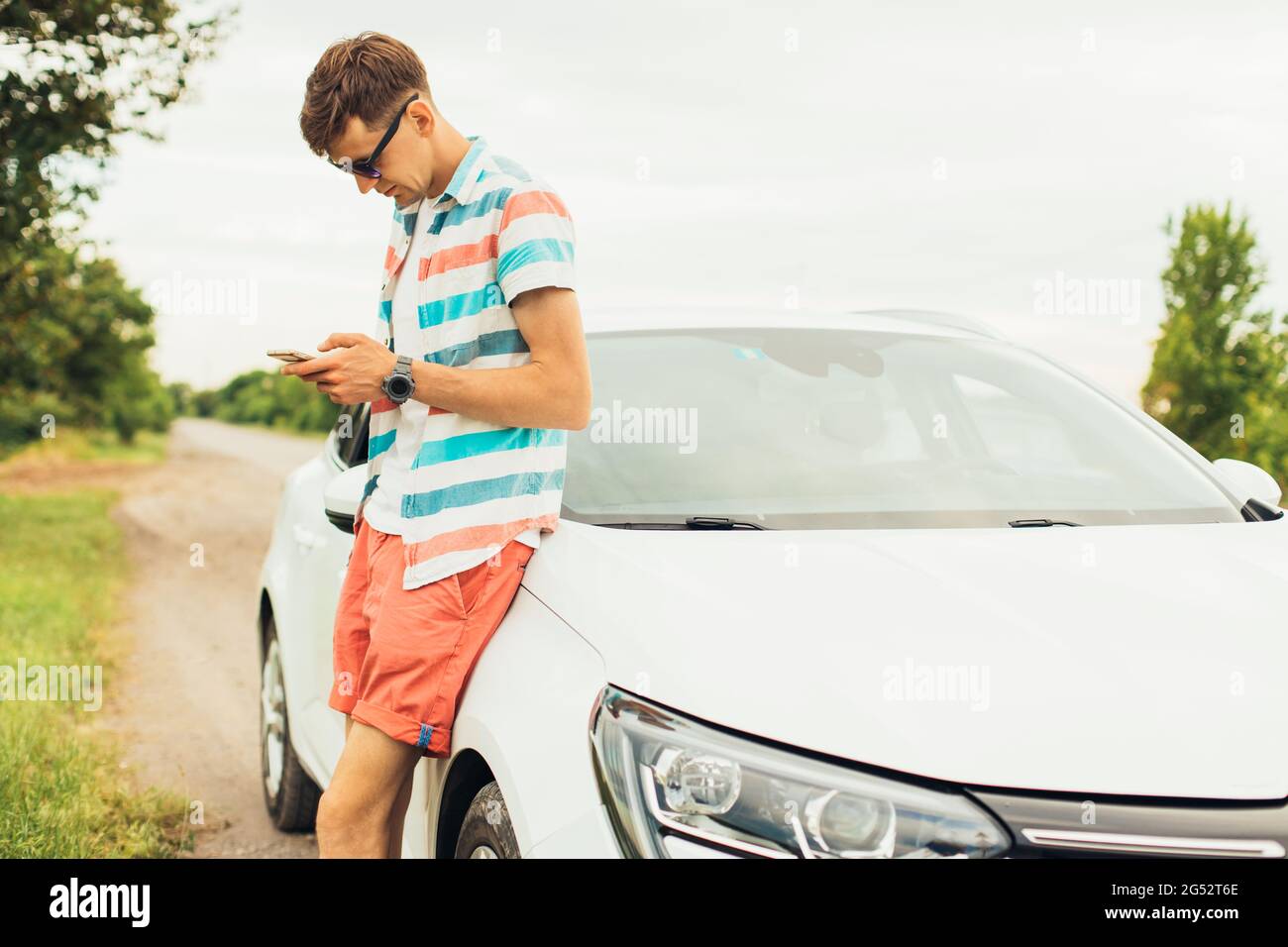 Hombre joven guapo en ropa de verano y gafas de sol de pie cerca de su coche blanco de lujo y utilizando smartphone, sosteniendo en la mano, al aire libre en un sol de verano Foto de stock