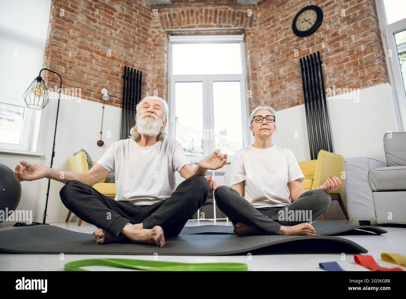 Dos personas mayores en activo sentados en posición de loto sobre  colchoneta de yoga y meditando con ojos cerrados. Ejercicios domésticos para  la salud mental. Formación familiar de edad avanzada en el