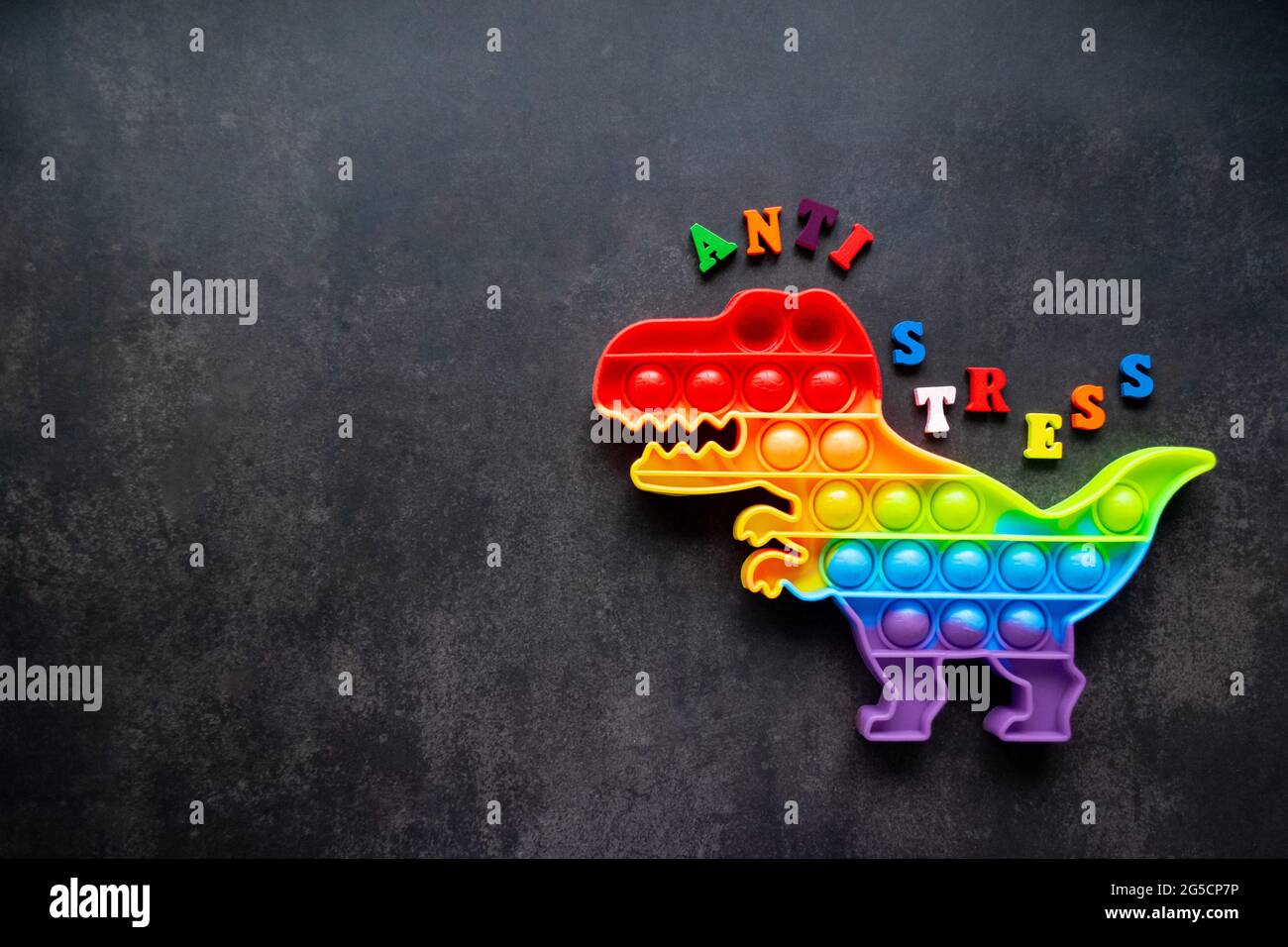 Juguete pop que dinosaurio colores arco iris sobre un fondo negro con letras multicolores y la inscripción-Anti-estrés en la espalda. Espacio de copia. Foto de stock
