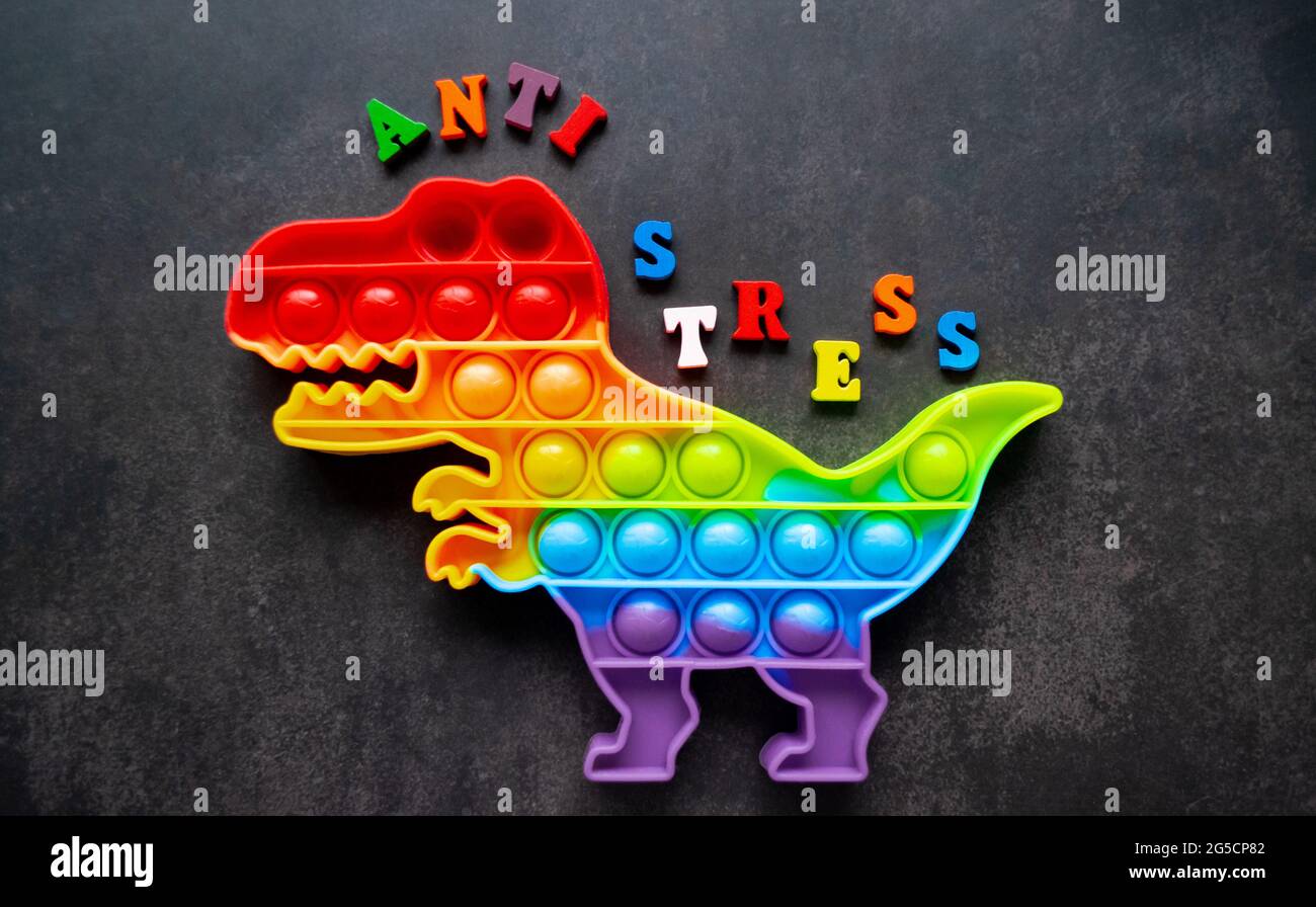 Juguete pop que dinosaurio colores arco iris sobre un fondo negro con letras multicolores y la inscripción-Anti-estrés en la espalda. Foto de stock