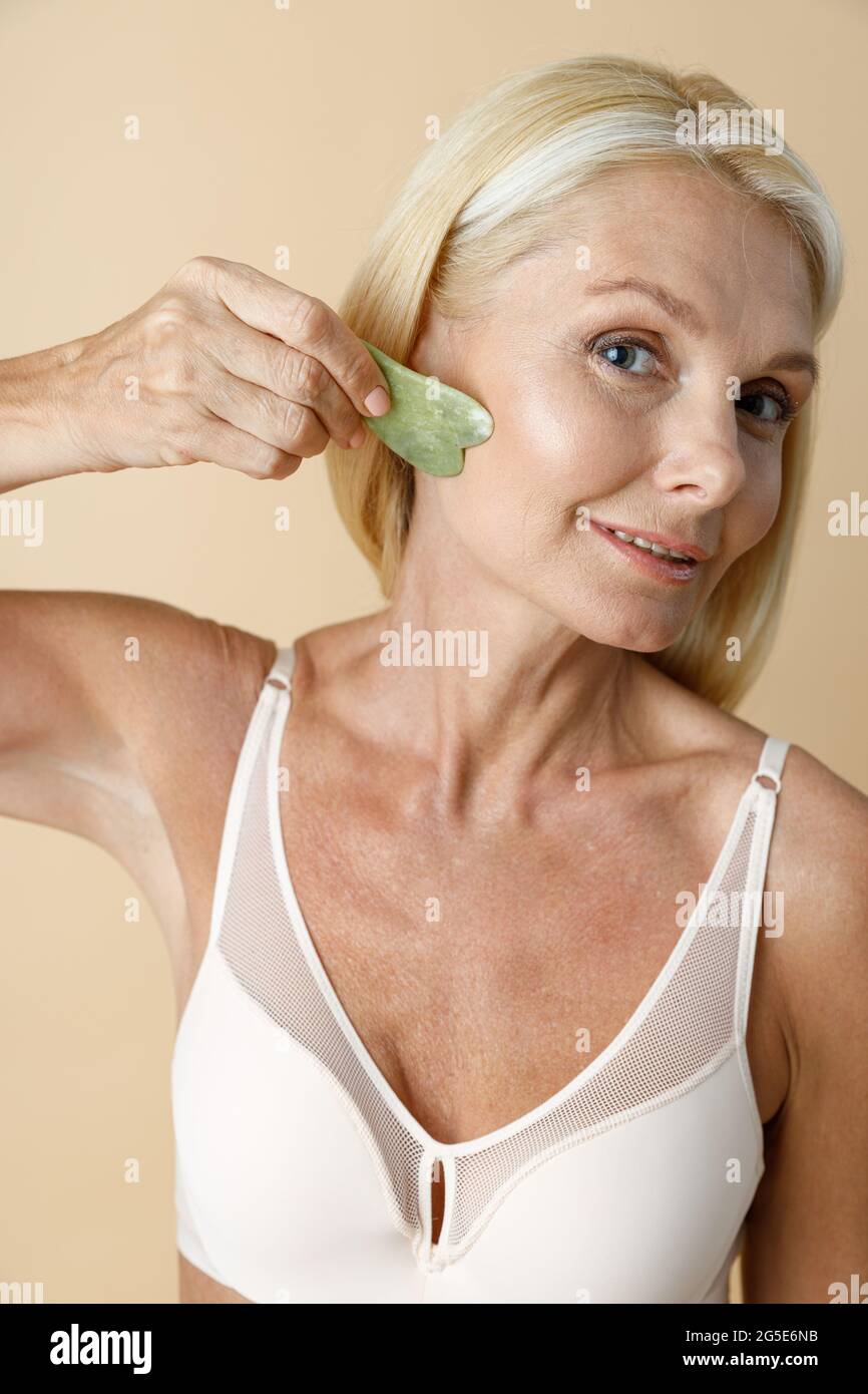 mujer rubia madura en sujetador blanco la herramienta de masaje jade Gua Sha para el cuidado de la piel y el tratamiento de belleza aislado sobre fondo beige Fotografía de