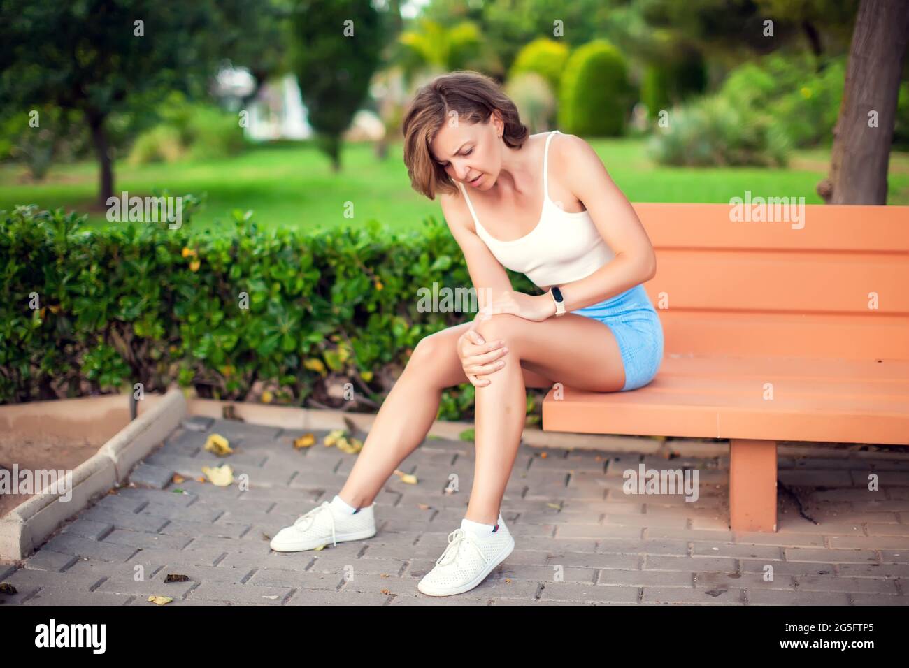 Dolor de rodilla. Mujer con lesión en la rodilla sentada en el banco al aire libre. Concepto de salud y estilo de vida Foto de stock