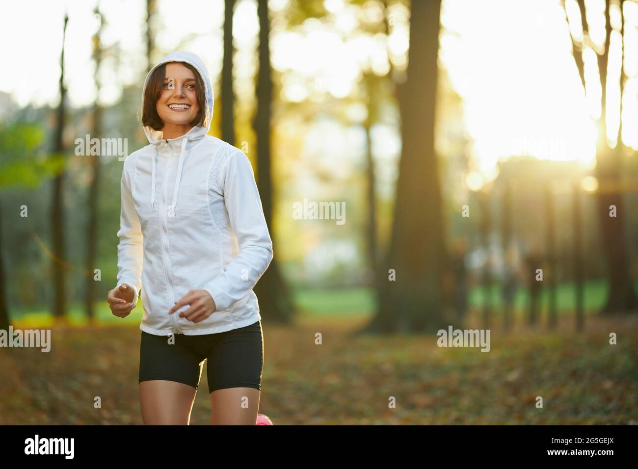 Sonriente mujer de fitness con chaqueta blanca con capucha haciendo  ejercicio por la mañana en el aire fresco. Mujer activa en ropa deportiva  haciendo ejercicio regular para mantenerse en forma y saludable