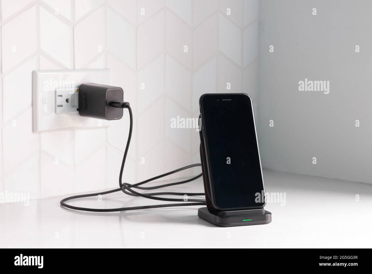 un teléfono inteligente negro conectado a una estación de carga inalámbrica conectado a una encimera de cocina o banco blanco con espacio para copias Foto de stock