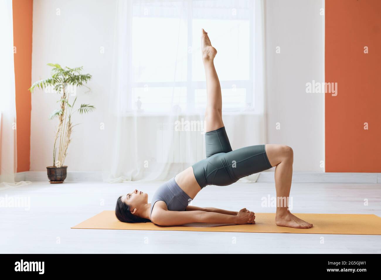 Mujer joven en leggings y camiseta corta, practicando yoga, haciendo dvipada pithasana con la pierna extendida hacia arriba, pose de medio puente Foto de stock