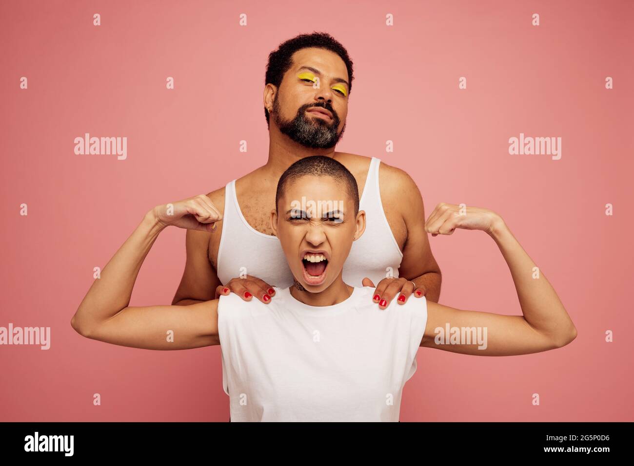 Retrato de amigos expresivos sobre fondo rosa. Mujer con cabeza afeitada flexionando sus bíceps y hombre barbudo con maquillaje detrás de ella. Foto de stock