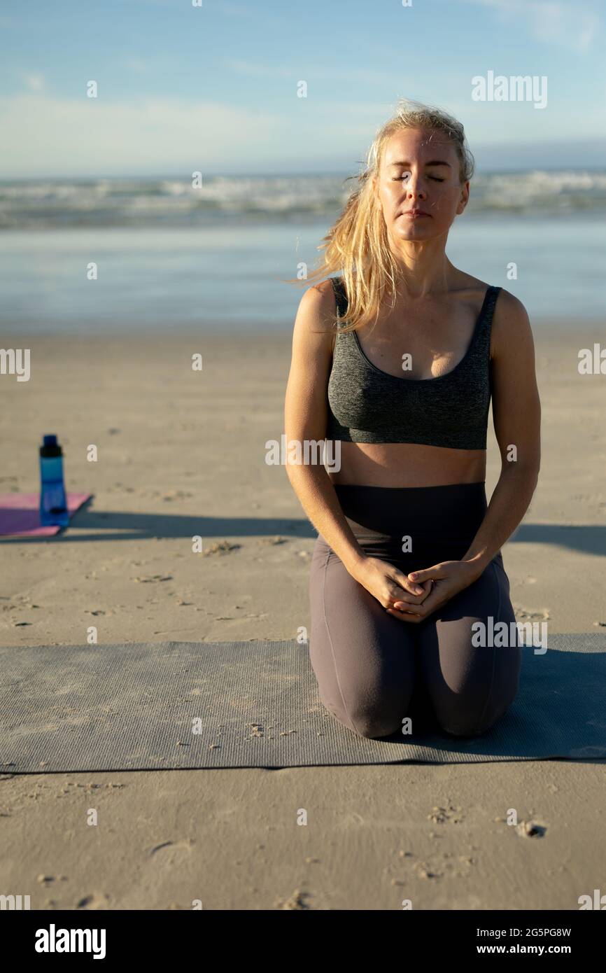 Mujer caucásica practicando yoga, arrodillándose meditando en la playa Foto de stock