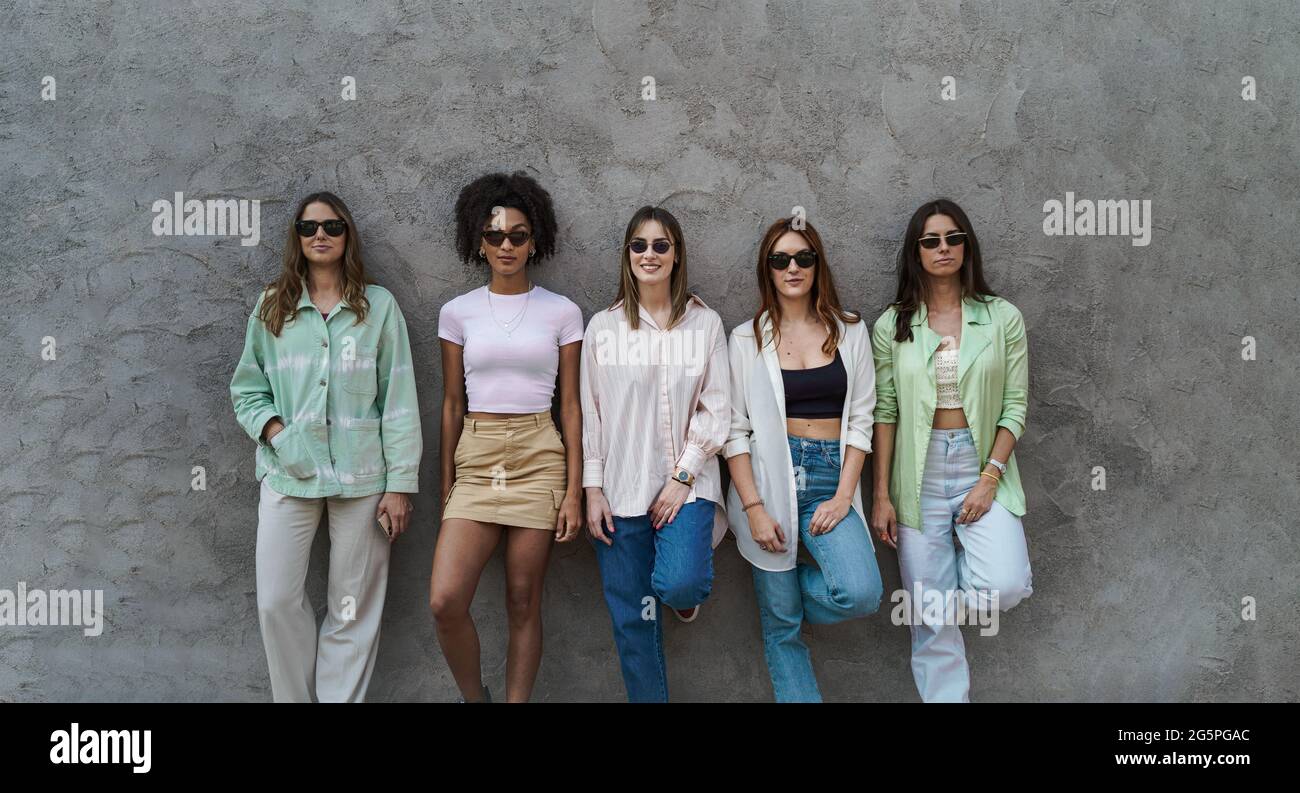 Retrato de cinco chicas de pie en una pared de hormigón divirtiéndose, usando gafas de sol y viendo la cámara. Concepto de niñas de la generación del milenio solamente. Foto de stock