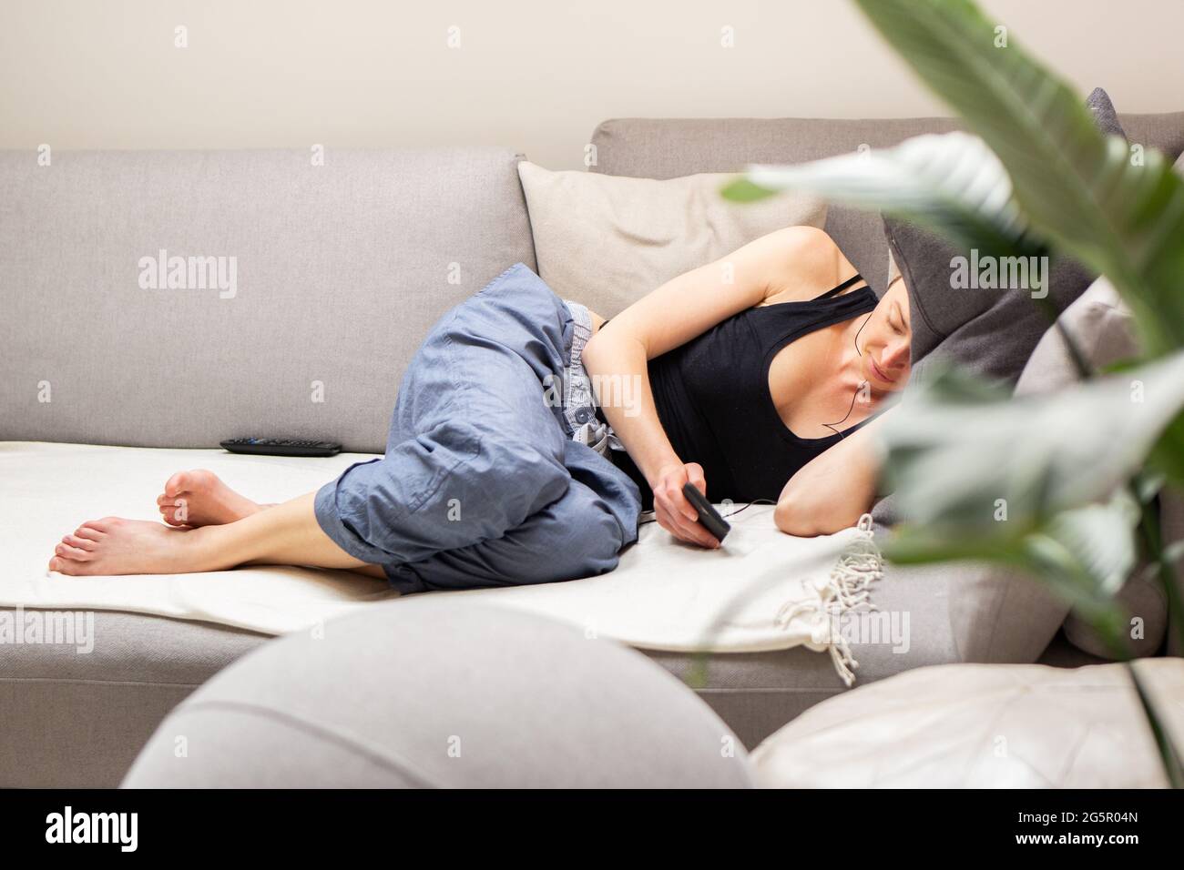 Mujer joven cansada, descansando, tumbada en un cómodo sofá con grandes cojines en la sala de estar del hogar utilizando su teléfono móvil. Foto de stock