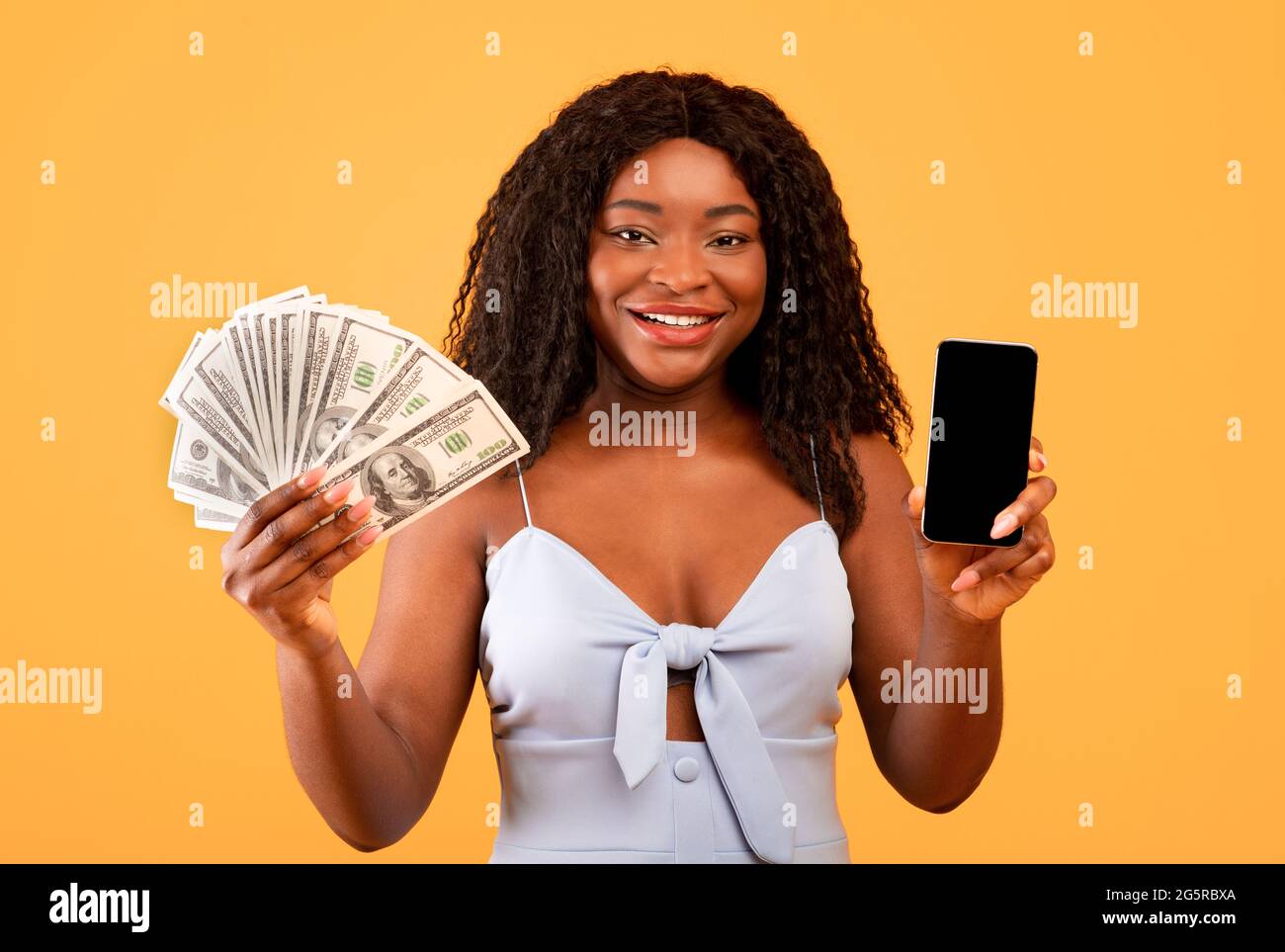 Aplicación de compras en línea. Mujer negra que muestra el smartphone con pantalla en blanco, que sostiene el ventilador del dinero, la publicidad de la nueva tienda web Foto de stock