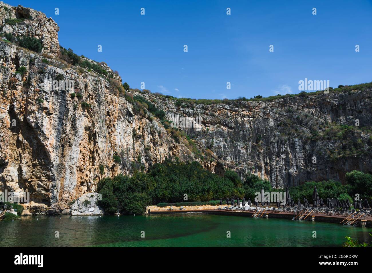 Lago Vouliagmeni en Grecia en el soleado día de verano. Pequeño lago salobre alimentado. Paisaje escénico de montaña blanca y agua verde. Muelle térmico. SPA Foto de stock