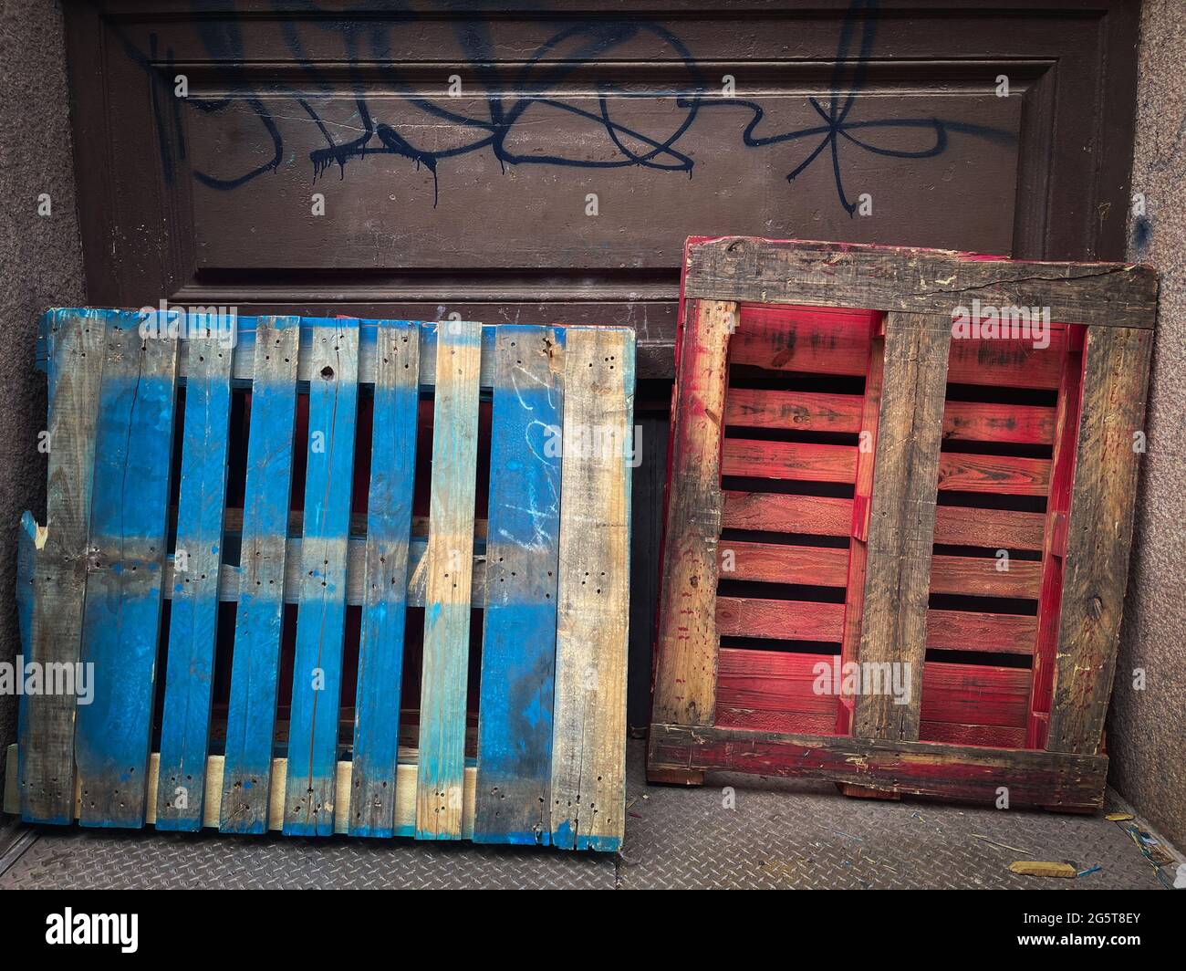 Palets de madera de colores pintados de azul y rojo apoyado contra una puerta de garaje de metal con graffiti negro escrito en nit Foto de stock