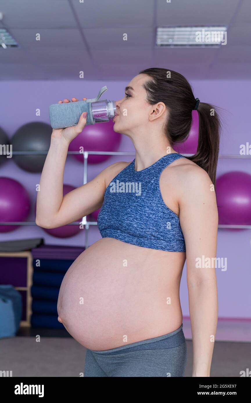 Una mujer embarazada en ropa deportiva bebe agua en el gimnasio con espadas de fitness. La niña se dedica a Pilates para las mujeres embarazadas. Último trimestre Fotografía de stock -