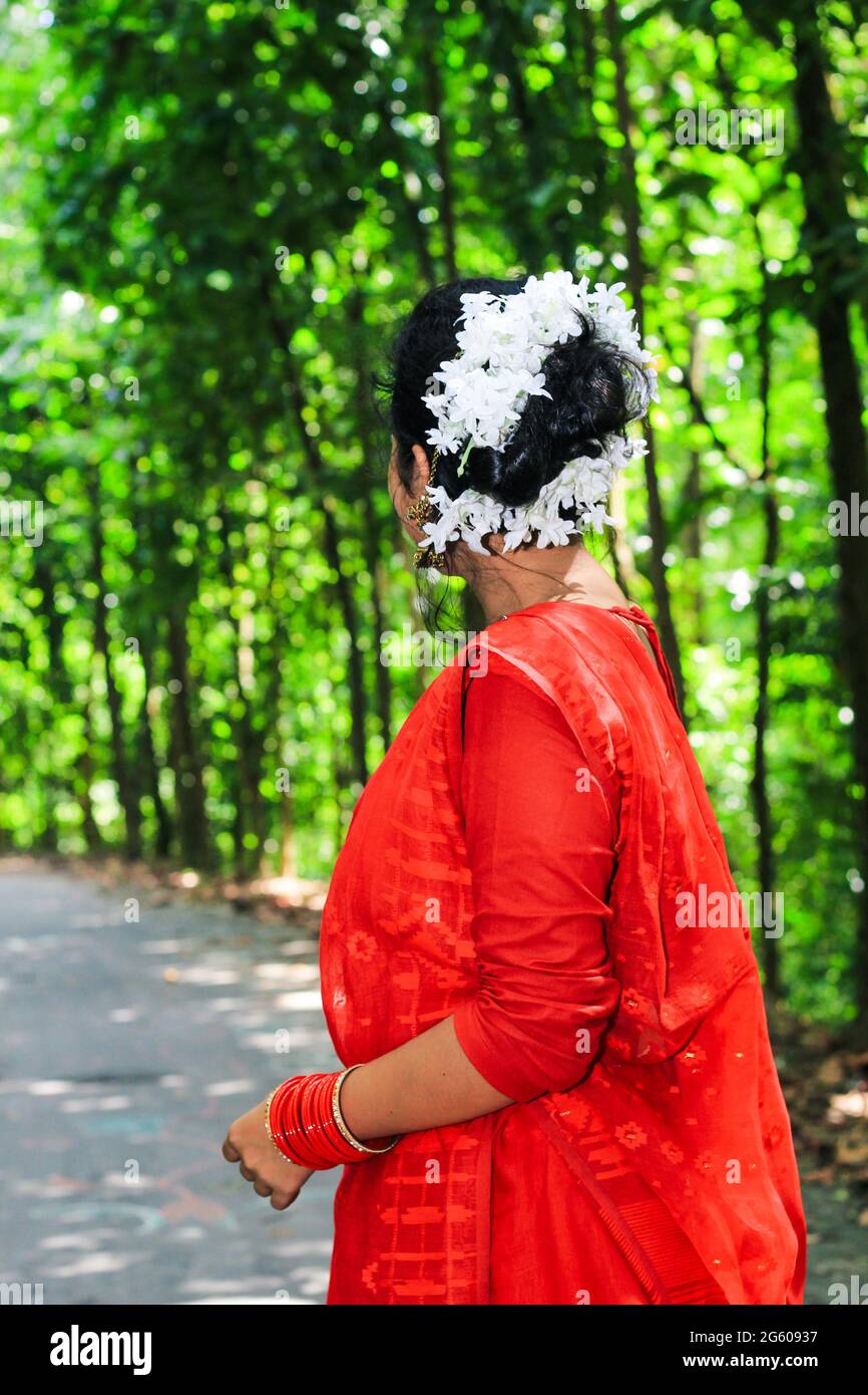 Mujer joven vistiendo un sari rojo caminando sobre camino verde asfalto. La chica camina a lo largo del camino en el bosque a la luz. Foto de stock