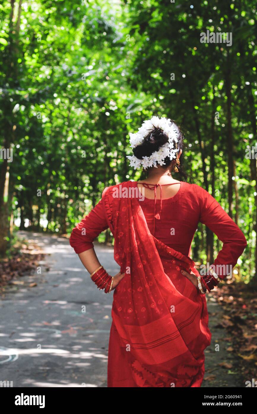 Mujer joven vistiendo un sari rojo caminando sobre camino verde asfalto. La chica camina a lo largo del camino en el bosque a la luz. Foto de stock