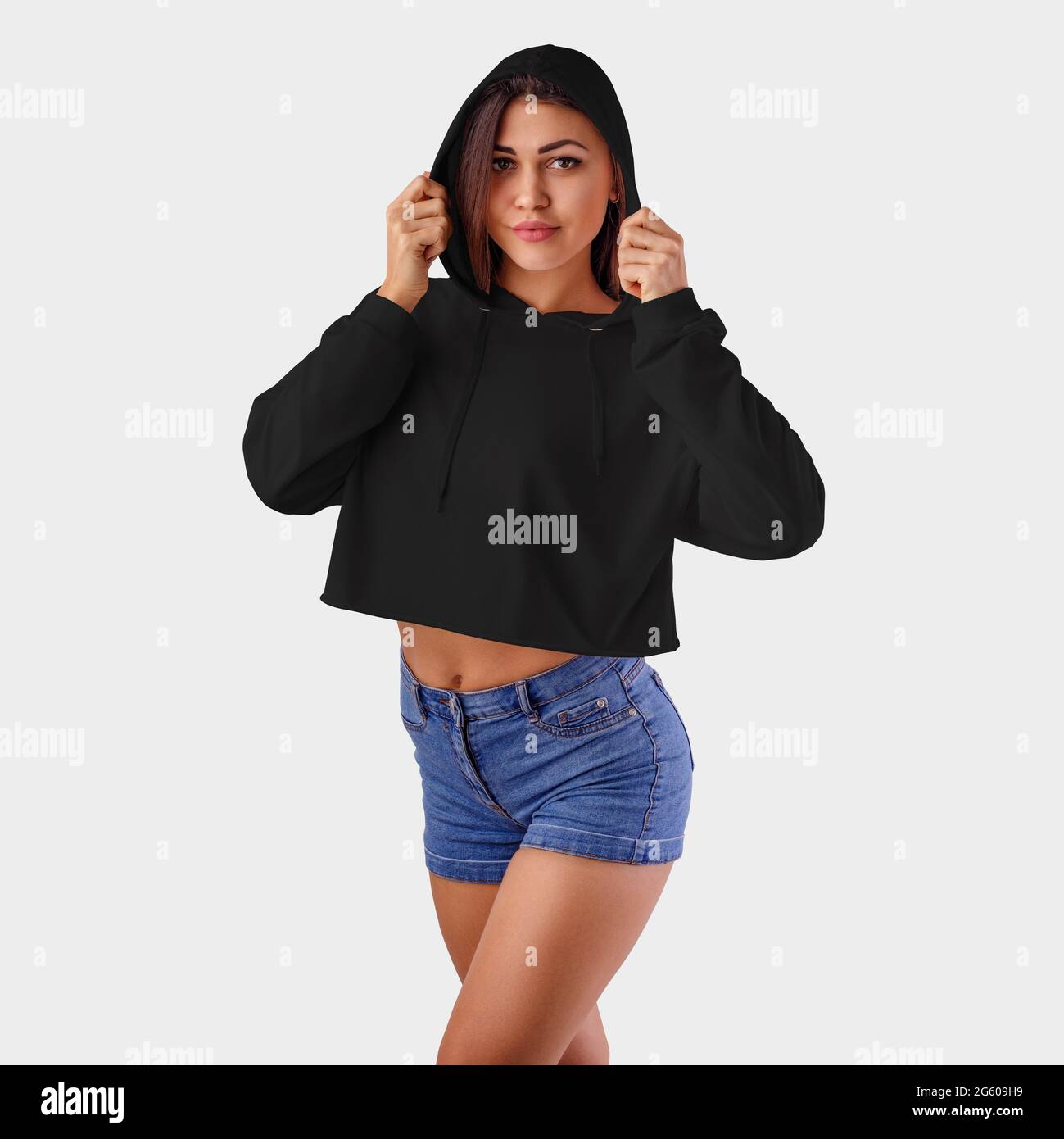 Burlándose de una camiseta corta hembra negra sobre una chica bonita en  pantalones cortos azules, usando una capucha, en el fondo en el estudio.  Plantilla con capucha en blanco para presenta Fotografía