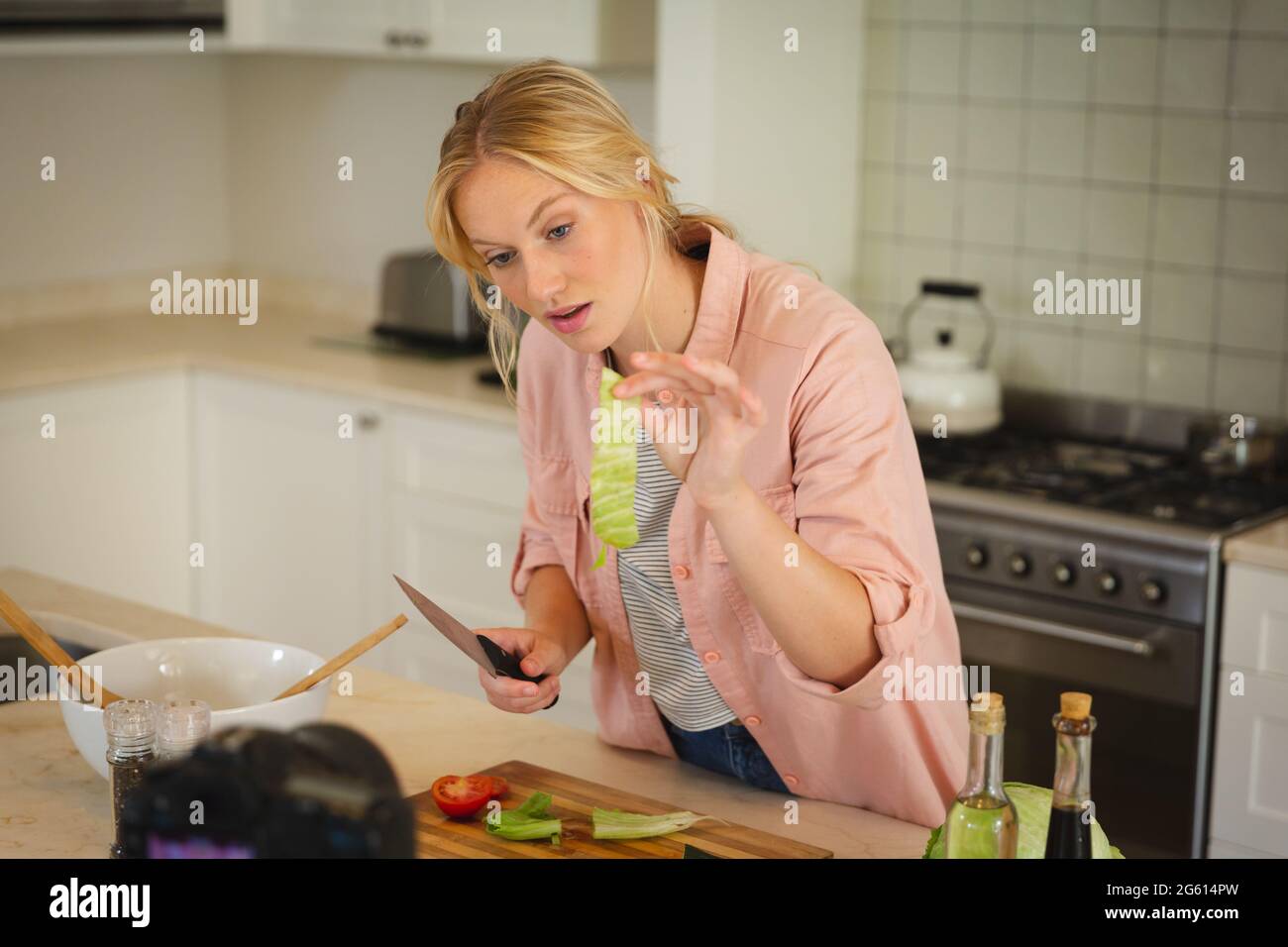 Mujer caucásica en la cocina sosteniendo lechuga y hablando con la cámara, haciendo vlog cocinar Foto de stock