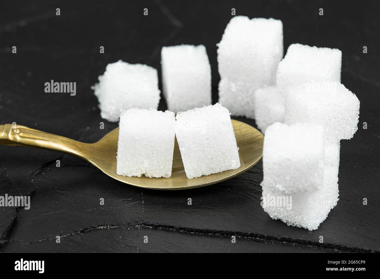 Se colocaron varios cubos de azúcar blanco en una cuchara de latón sobre un fondo de piedra negra. Foto de stock