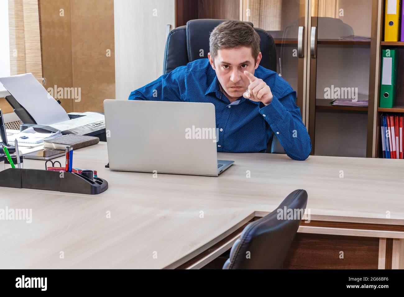 Un jefe enojado sentado en una mesa con un ordenador portátil apunta con el dedo a un subordinado. Una severa reprimenda al subordinado. Trabajador de oficina. Gerente del departamento Foto de stock