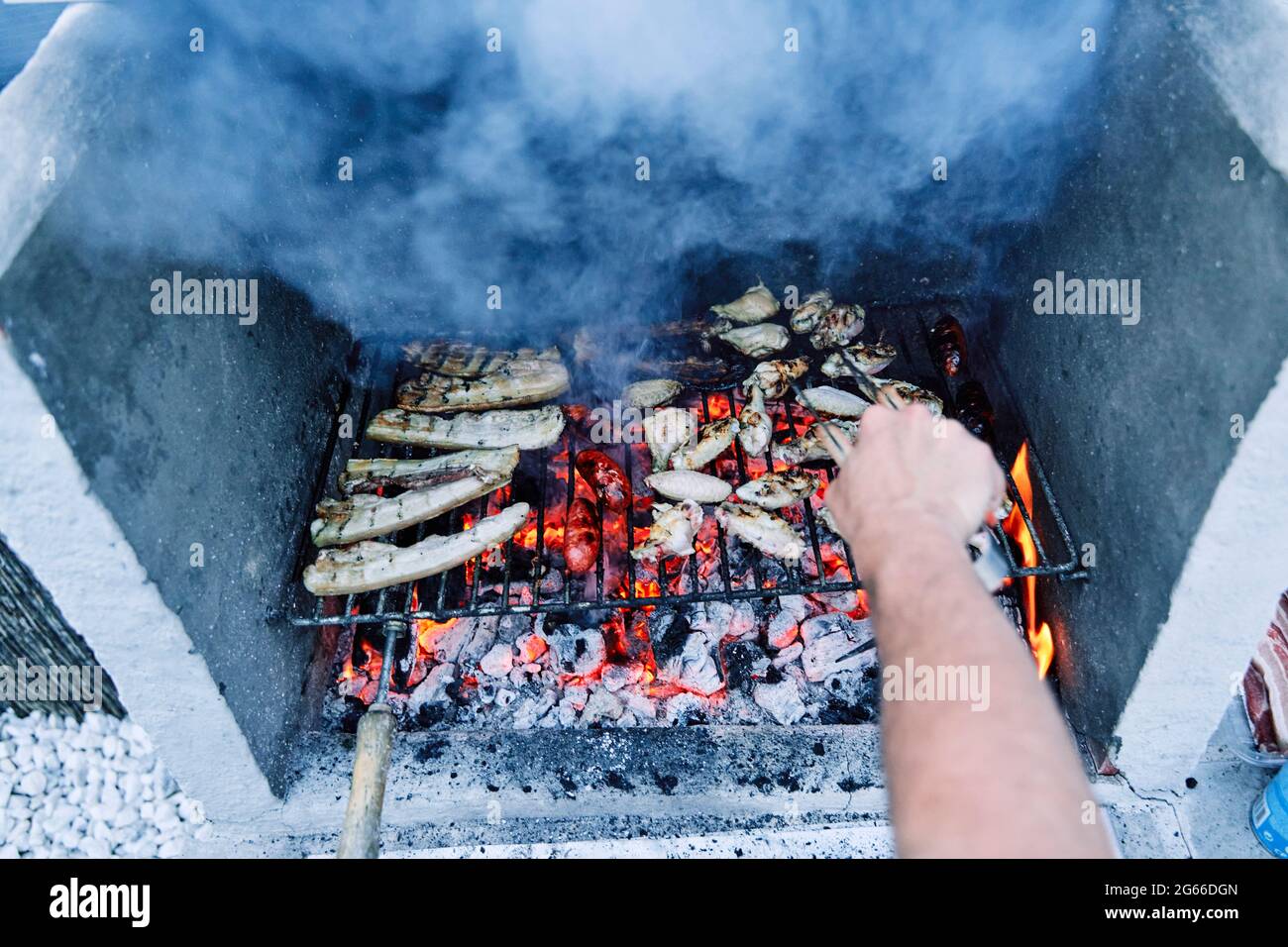 Detalle de la mano de un hombre con pinzas que se convierte en deliciosa carne a la parrilla sobre los carbones de una barbacoa. Preparar la cena al aire libre. Foto de stock