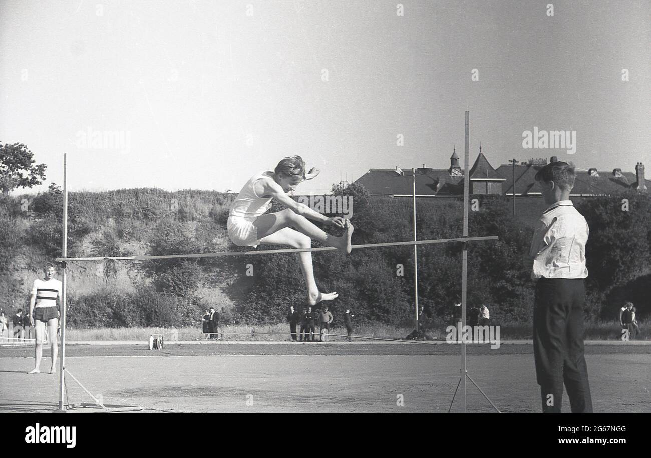 1960s, histórico, atletismo, deportes de escuela inter-condado, un  estudiante haciendo el salto alto en los pies descalzos, utilizando la  técnica tradicional de tijeras, observado por otro niño, manteniendo su ojo  en el