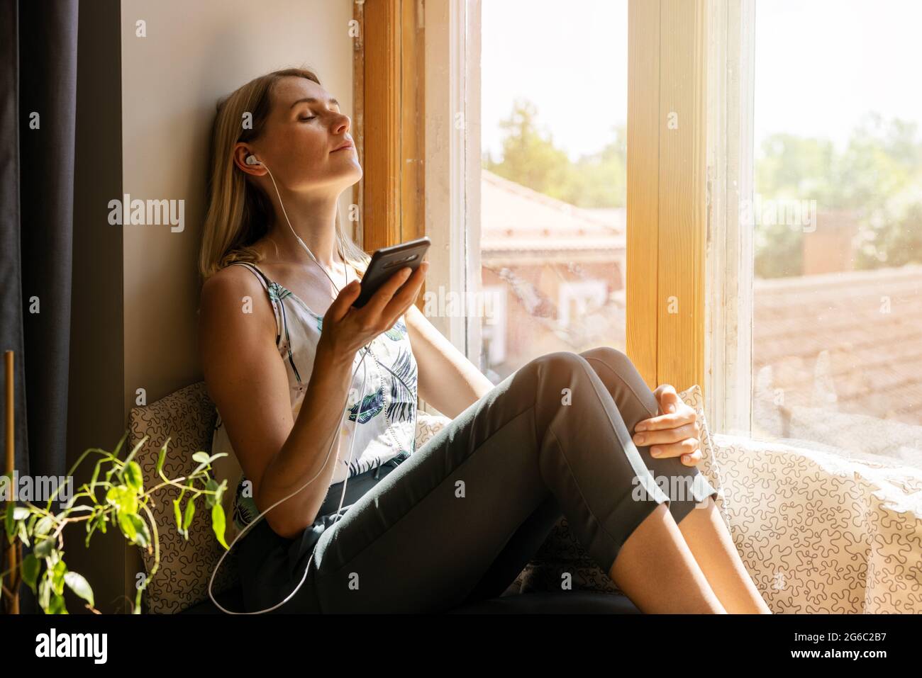 mujer relajándose en el soleado alféizar de la ventana con los ojos cerrados y escuchando música con los auriculares del smartphone Foto de stock