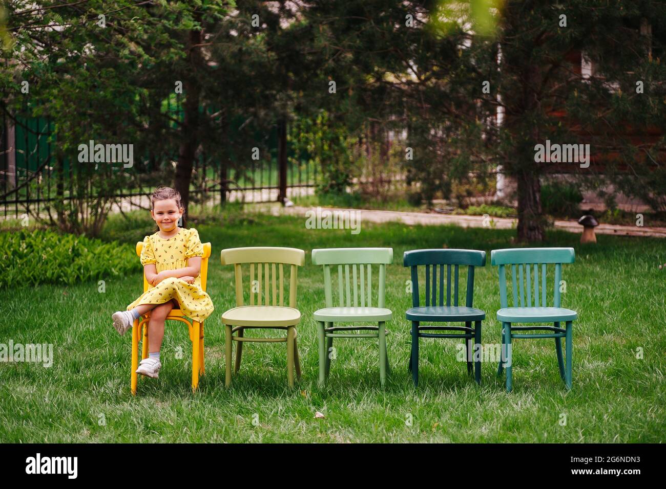 Una chica alegre y riendo con un vestido amarillo se sienta en sillas pintadas multicolores vienesas esperando a un amigo para una fiesta infantil y cumpleaños Foto de stock