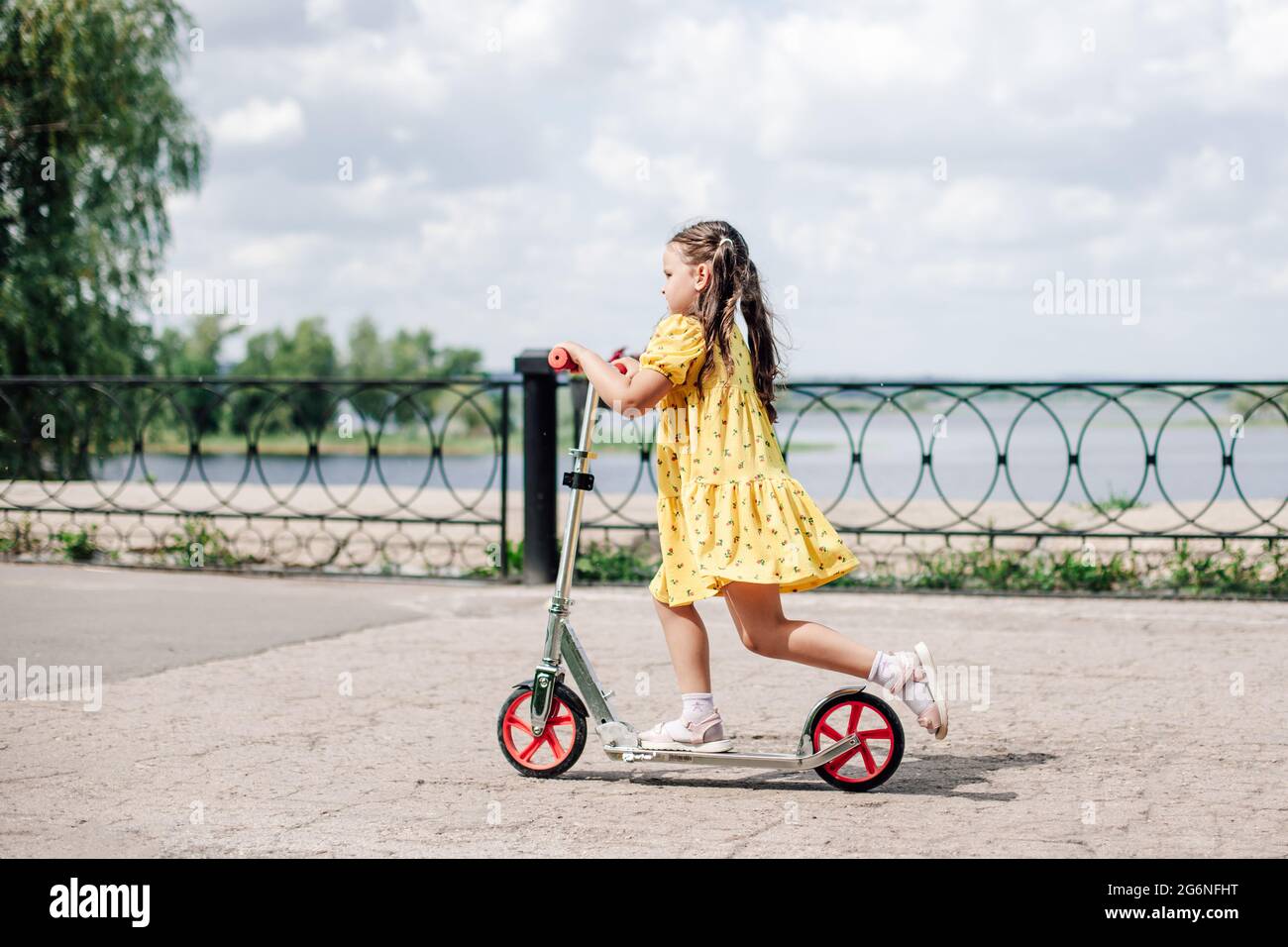 retrato estilo de vida de una niña de talla completa con un vestido  amarillo montando un scooter a lo largo de la orilla del lago, vacaciones  familiares en la naturaleza, unidad con