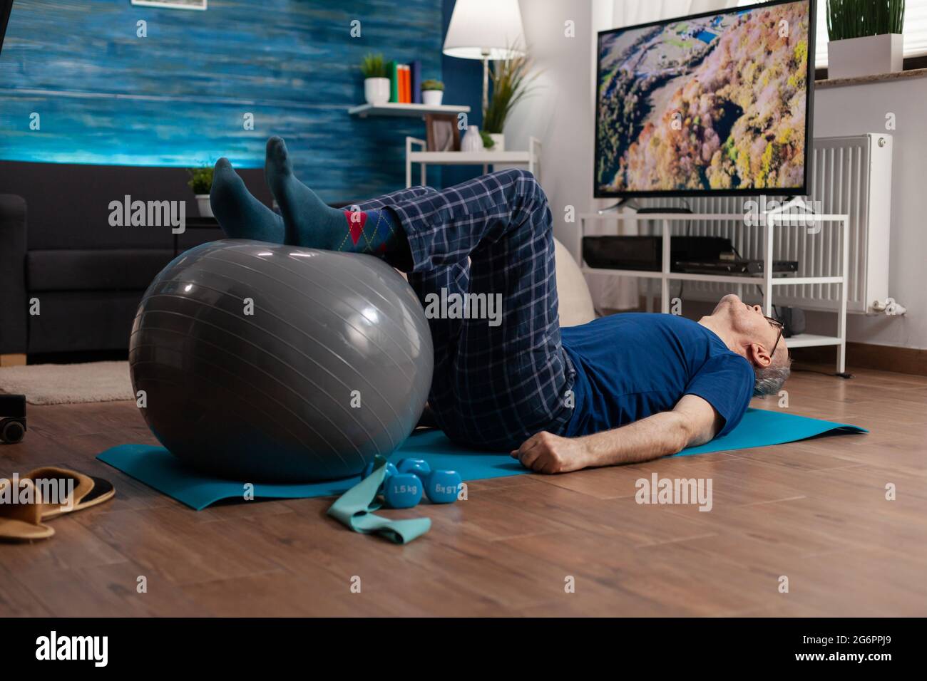 Jubilado hombre mayor haciendo bienestar calentar piernas con pelota suiza sentado en colchoneta de yoga en la sala de estar. Jubilado pensionista que practica ejercicios abdominales trabajando en la salud muscular del cuerpo Foto de stock