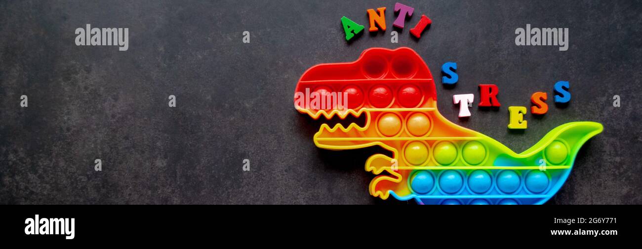 Bandera con juguete pop que dinosaurio colores arco iris sobre un fondo negro con letras multicolores y la inscripción anti-estrés en la espalda. Espacio de copia Foto de stock