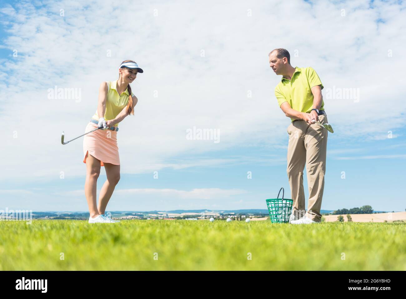 Longitud total de una joven mujer sonriendo mientras practica el movimiento correcto para golpear durante la clase de golf con un profesional experimentado jugador afuera Foto de stock