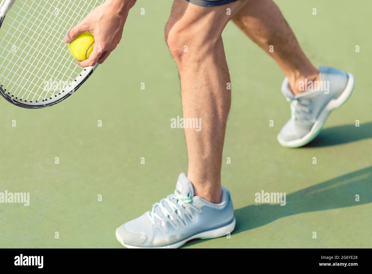 Sección baja de un jugador profesional que lleva calzado deportivo gris mientras sujeta la pelota y la raqueta de tenis durante el partido sobre una superficie verde Foto de stock