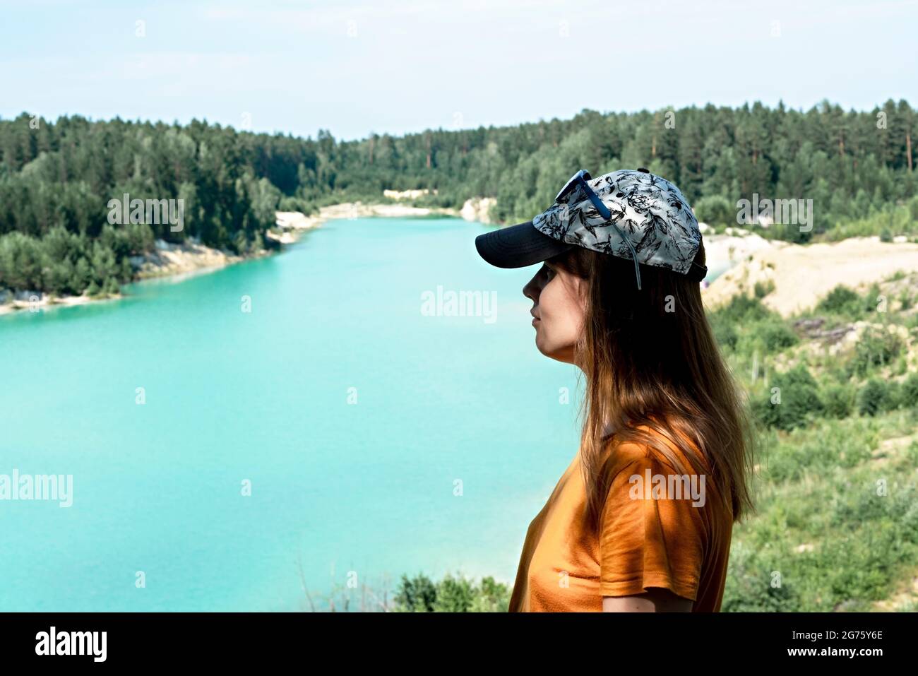 Vista trasera de una joven mujer rubia viajera en una camiseta de gorra y mostaza en el acantilado de color azul turquesa lago o río y bosque coníferas, tr Foto de stock