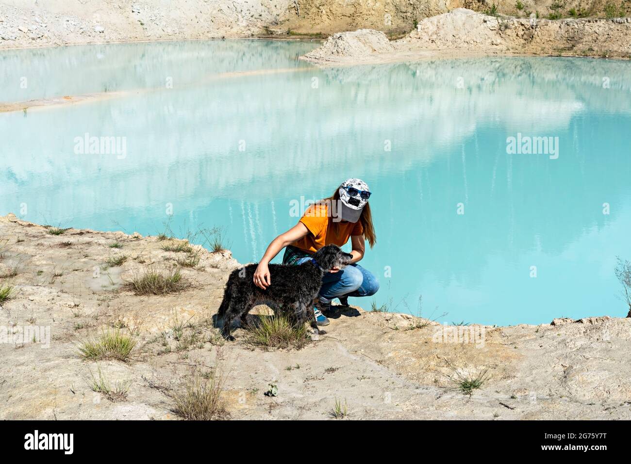 Vista trasera de una joven viajera con gorra y camiseta de jengibre acariciando un perro gris esponjoso en la orilla arenosa del lago o río azul, a las mascotas les encanta el estilo de vida activo, Foto de stock