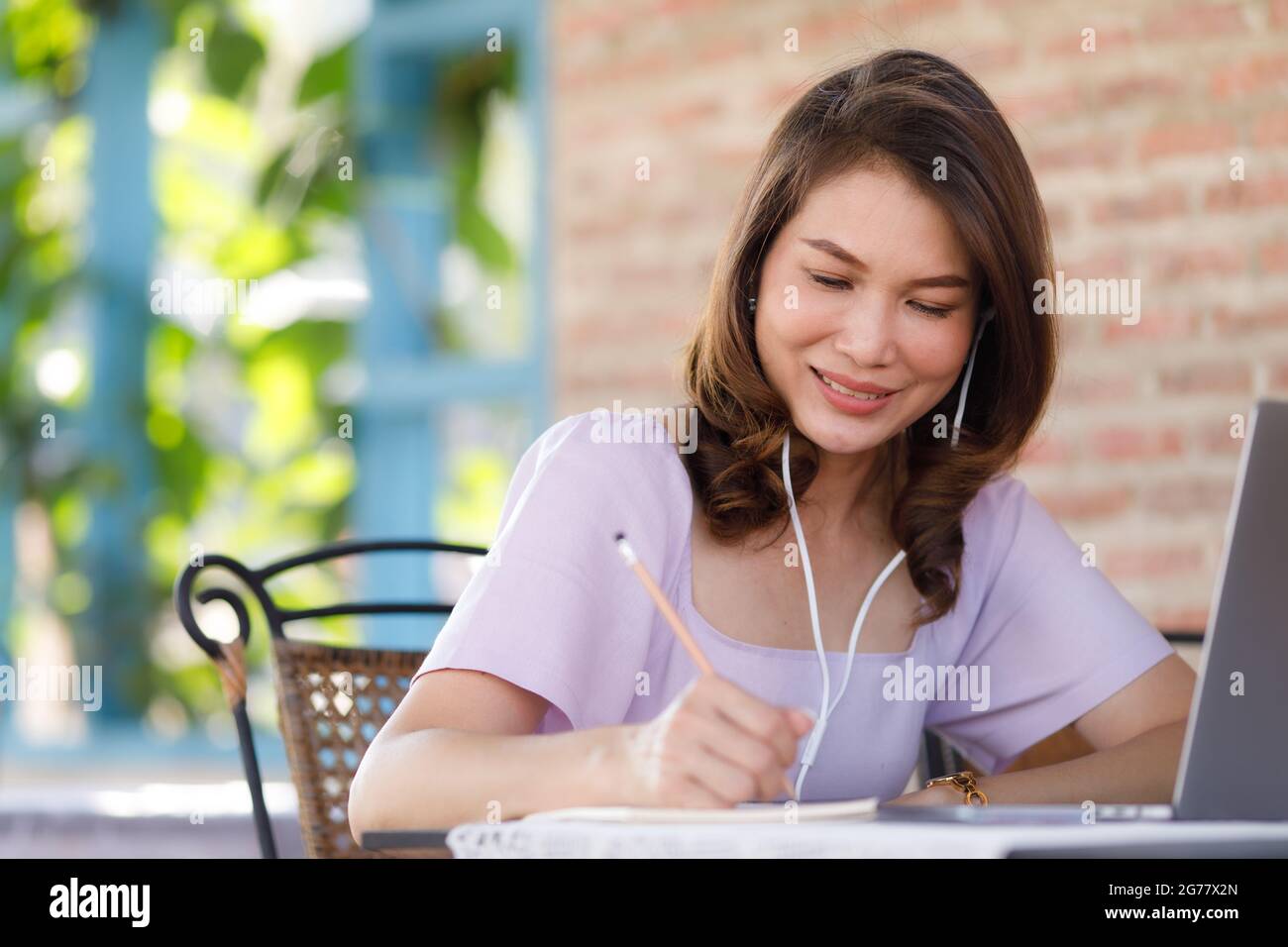 Hermosa mujer de mediana edad sentada en la tienda usando un lápiz para escribir en un cuaderno de papel con ordenador portátil y auriculares. W Fotografía de stock -