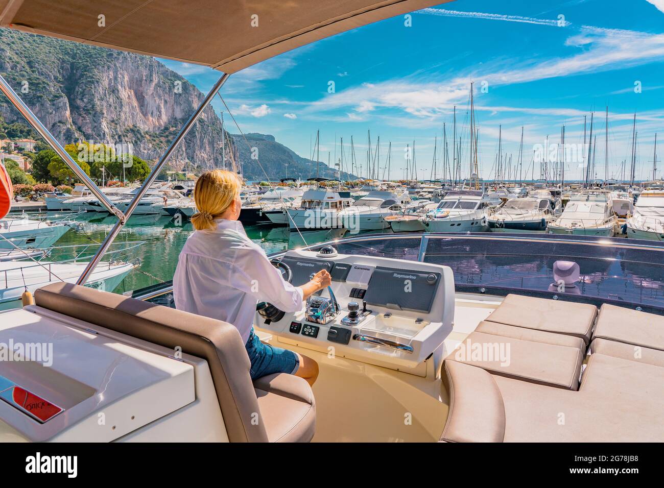 Navegación en el mar. Hermosa mujer rubia que conduce yate. Mujer adulta modelo en una camisa blanca sentada al volante del yate y comenzar a conducir un barco de lujo desde el puerto. Joven mujer que conduce yate. Foto de stock