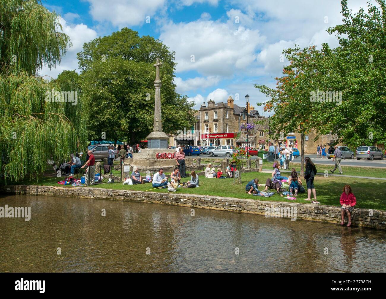 Reino Unido, GL Gloucestershire, Bourton-on-the-Water, gente sentada en las orillas del río Windrush bajo el sol, niños jugando. Bourton-on-the-Water es conocida como la Venecia de los Cotswolds. Aquí es donde comienza el Camino del Corazón de Inglaterra. Foto de stock