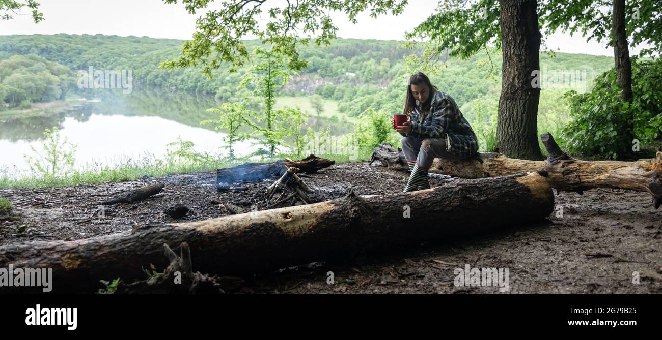 Una chica atractiva con una taza en su mano se sienta en un tronco y se calienta cerca de un fuego en el bosque. Foto de stock