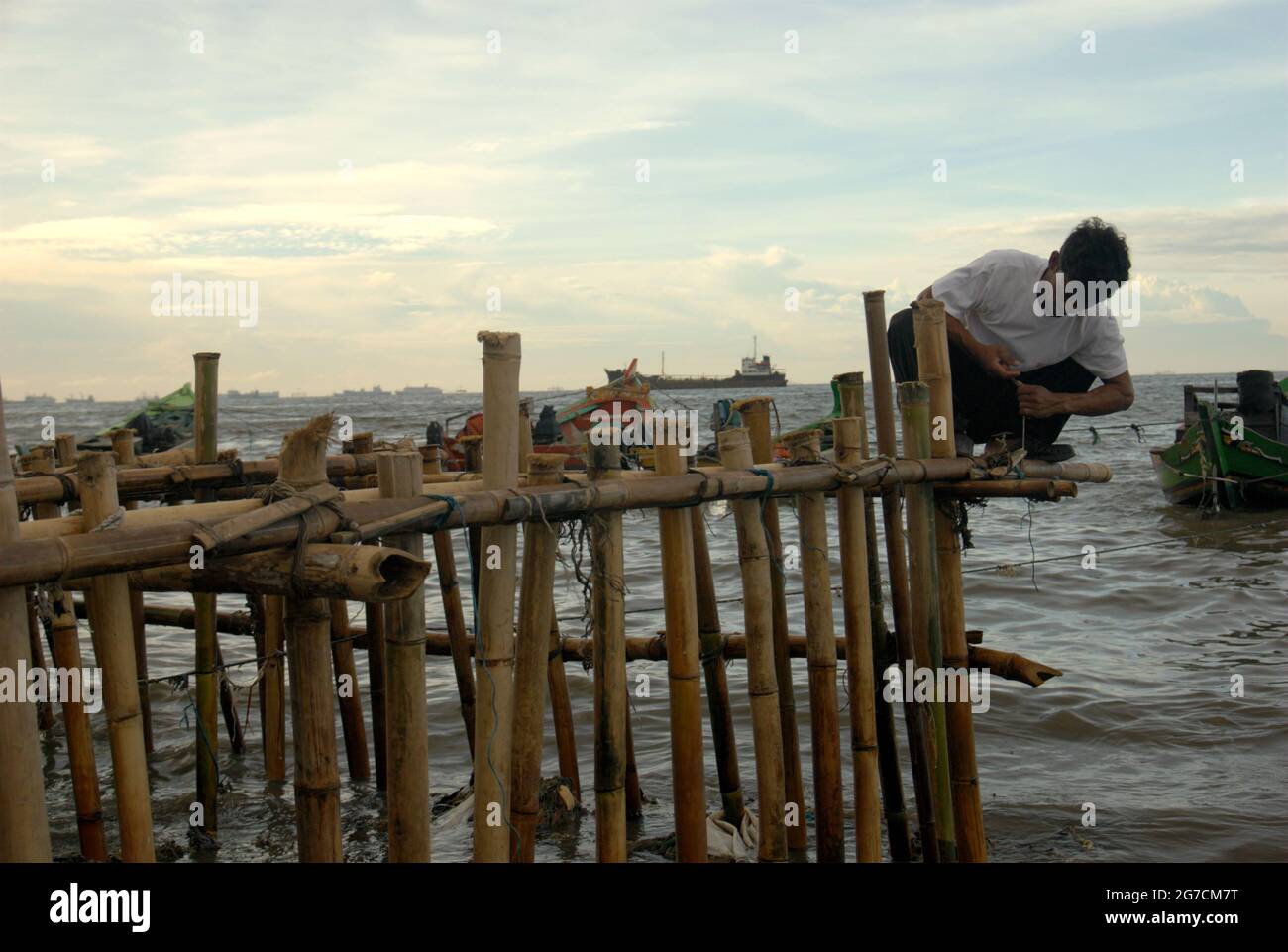 Yakarta, Indonesia. Un hombre está trabajando en una construcción de bambú sobre el agua costera en la aldea de pescadores de Cilincing, con la bahía de Yakarta y el puerto de Yakarta tráficos se pueden ver en la distancia. Foto de stock