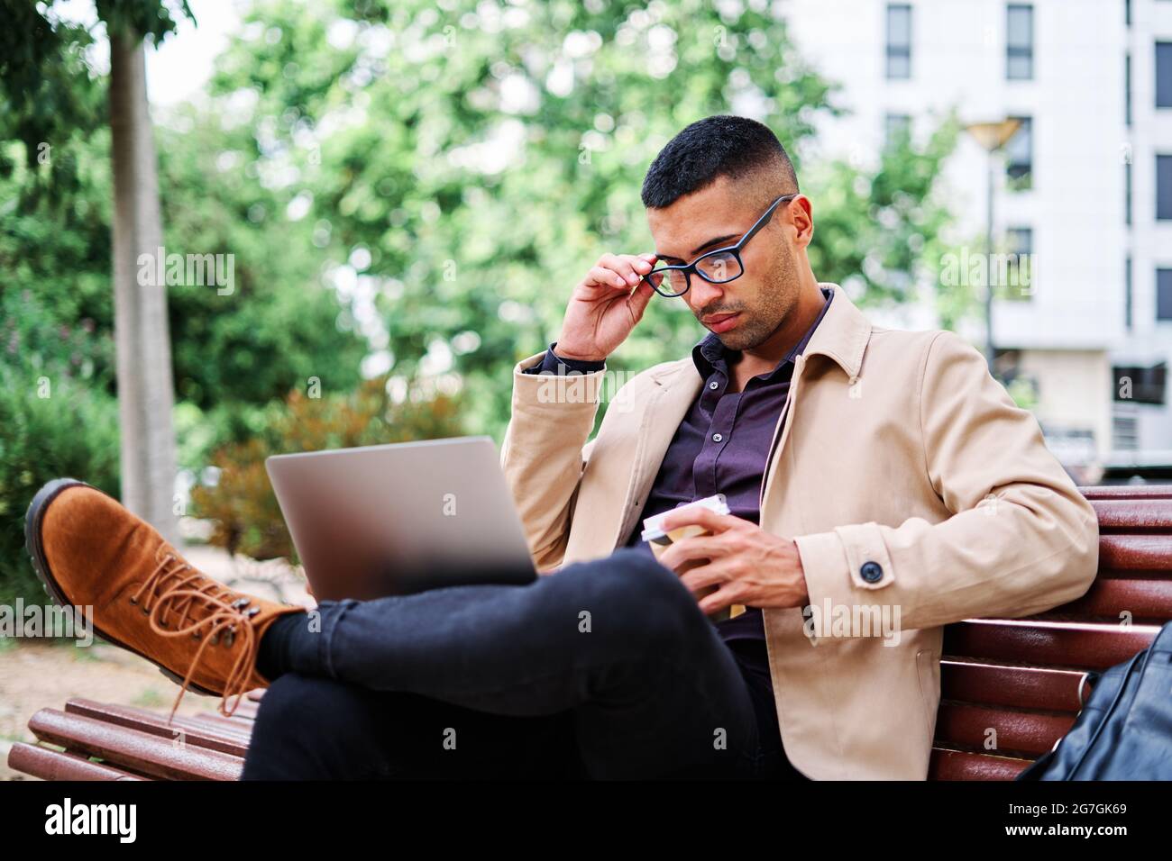 Largo de joven hispano confiado en con un traje elegante sport y gafas que  responden a la llamada mientras se sienta en el banco y trabaja con el  ordenador portátil o Fotografía