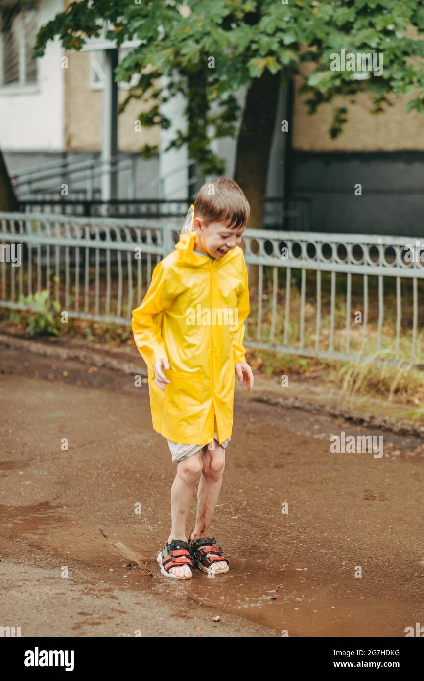 Un niño en un impermeable amarillo camina afuera bajo la lluvia. Un niño  salta en los nargues. Salte a través de charcos fangosos. Infancia feliz y  sin preocupaciones Fotografía de stock -