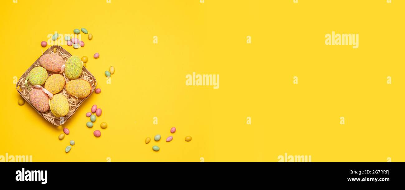 Cesta festiva con huevos de Pascua de colores y sabrosas habas de gelatina de caramelo de colores sobre un fondo amarillo con espacio para copias, vista superior, puesta plana. Horizontal Foto de stock
