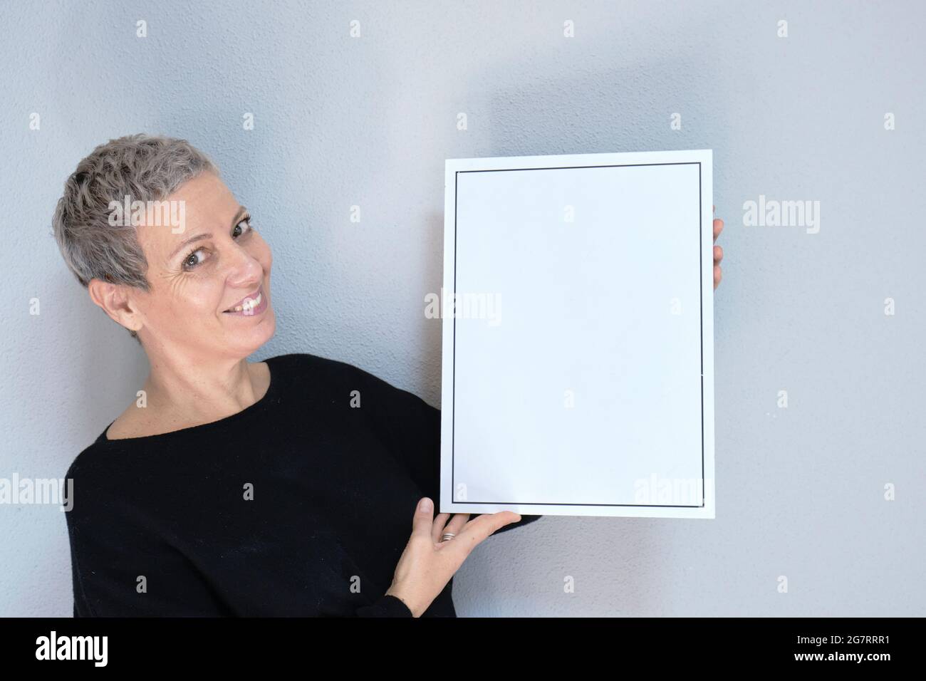 Retrato de primer plano de una mujer blanca adulta sonriente y atractiva con pelo gris mirando la cámara sosteniendo un marco vacío. Vista aislada sobre fondo gris Foto de stock