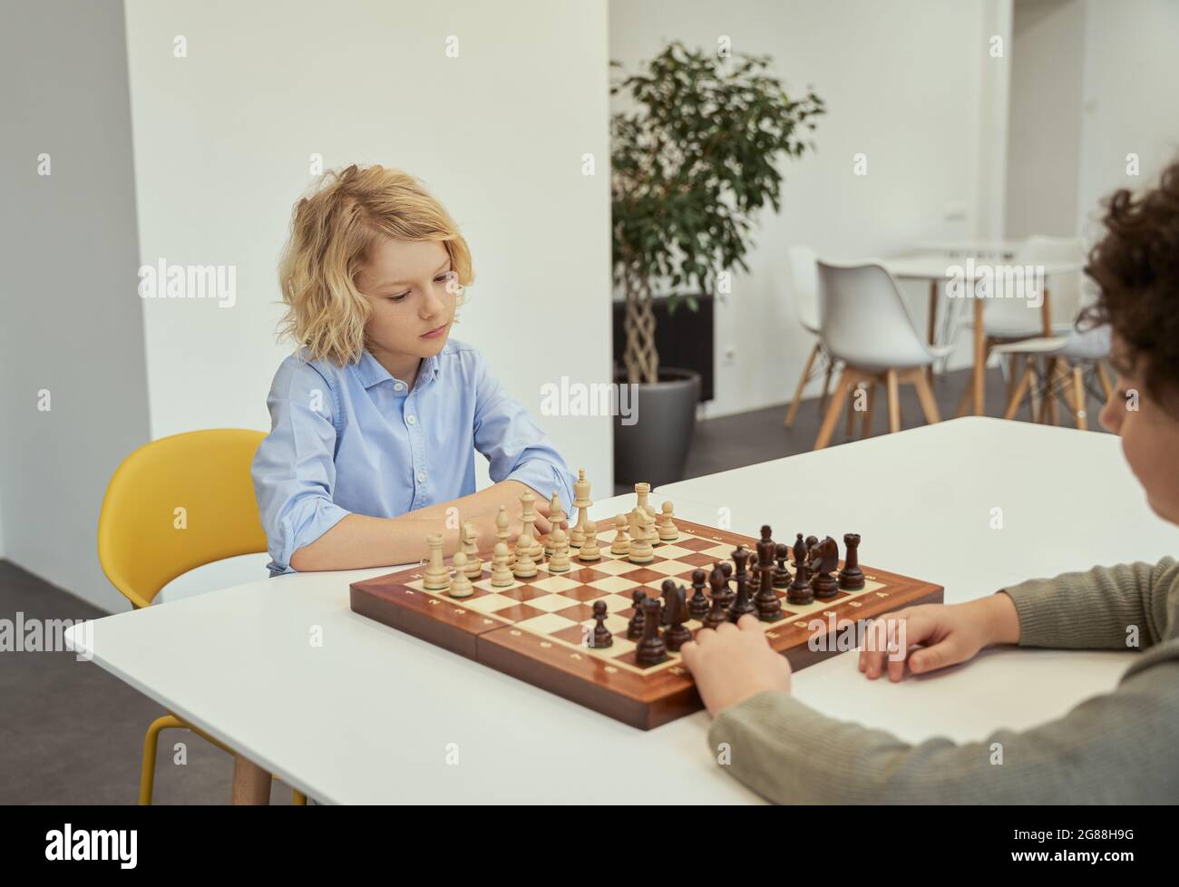 Piense antes de actuar. Un niño caucásico reflexivo jugando al ajedrez con su amigo, sentados juntos en la mesa de la escuela Foto de stock