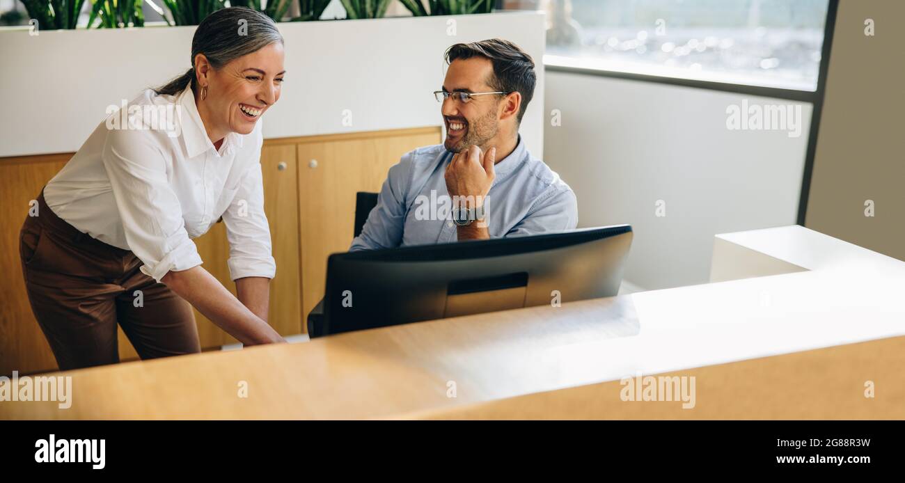 Feliz gerente hablando con un compañero sentado en el escritorio. Mujer madura ayudando al joven en el trabajo en la computadora en el escritorio de la oficina. Foto de stock