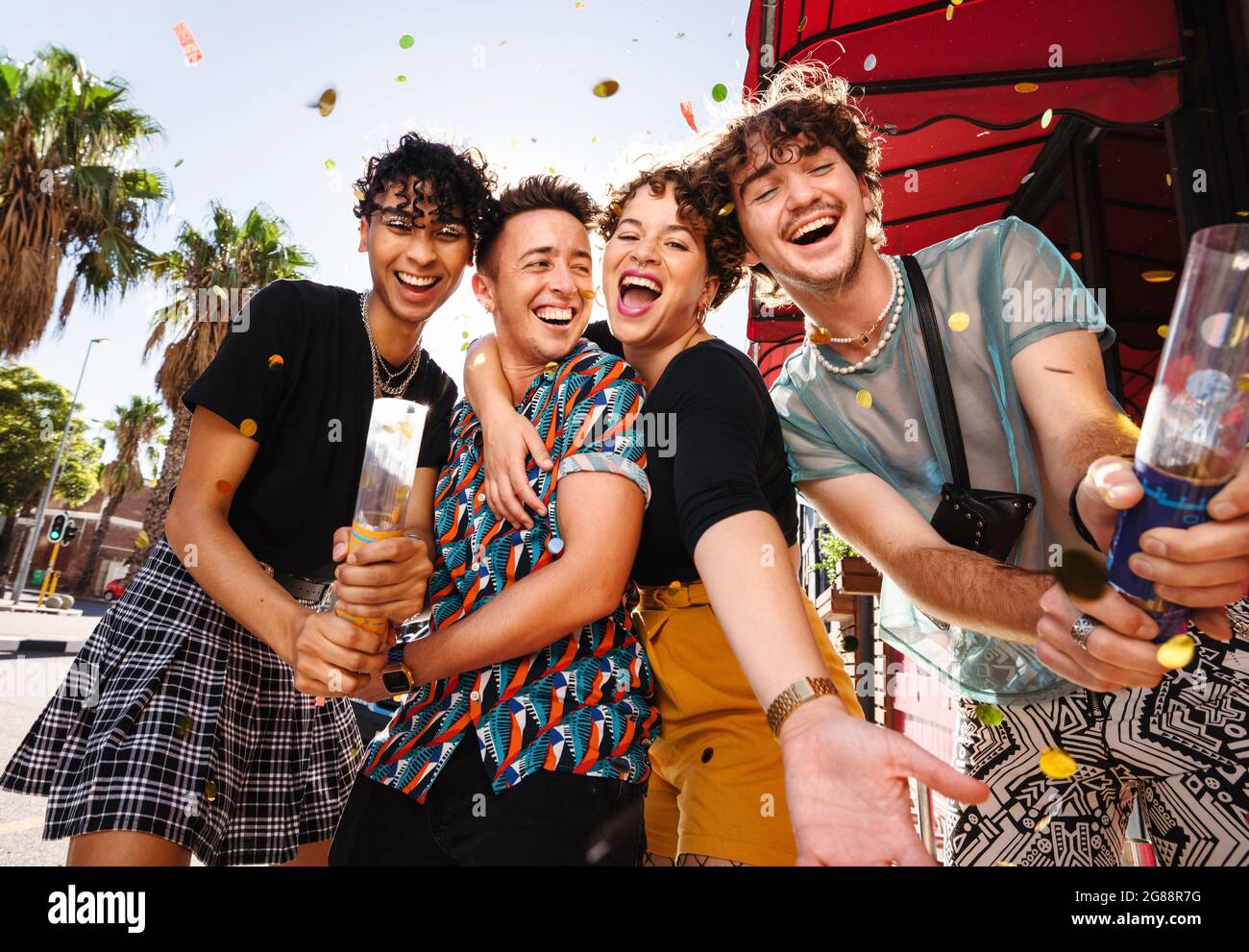 Cuatro amigos celebrando con confeti al aire libre. Grupo de gente queer feliz sonriendo alegremente mientras celebraba su amistad. Amigos que enlazan un Foto de stock